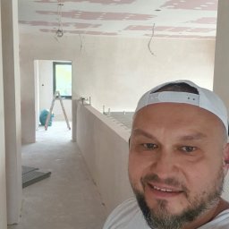 Drapinski Piotr Drapinski Usługi Ogólnobudowlane - Mężczyzna w czapce robi selfie na tle wnętrza w trakcie szpachlowania sufitu, widoczne kable i drabina w oddali.