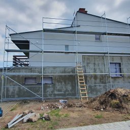 Drapinski Piotr Drapinski Usługi Ogólnobudowlane - Dom w trakcie prac elewacyjnych: szare ściany, rusztowania, zabezpieczone okna folią, sterta ziemi i gruzu u podstawy, drewniana drabina oparta o rusztowanie.