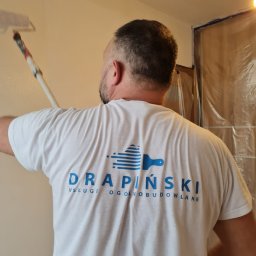 Drapinski Piotr Drapinski Usługi Ogólnobudowlane - Mężczyzna maluje ścianę wałkiem, ubrany w białą koszulkę z logo firmy budowlanej DRAPIŃSKI USŁUGI OGÓLNOBUDOWLANE, zabezpieczone folią fragmenty pomieszczenia.