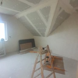 Drapinski Piotr Drapinski Usługi Ogólnobudowlane - Poddasze w trakcie montażu płyt kartonowo-gipsowych na skośnym suficie, widoczna drewniana drabina, płyty na podłodze, surowe ściany i okno z lewej strony.