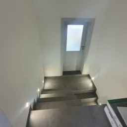 Nowoczesne schody wewnętrzne wyłożone szarą płytką, z oświetleniem punktowym w ścianie, prowadzące do białych drzwi z przeszkleniem.