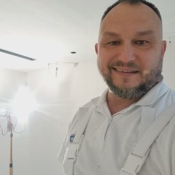 Drapinski Piotr Drapinski Usługi Ogólnobudowlane - Uśmiechnięty malarz w białym kombinezonie na tle świeżo pomalowanego, jasnego pomieszczenia z widoczną lampą na statywie.