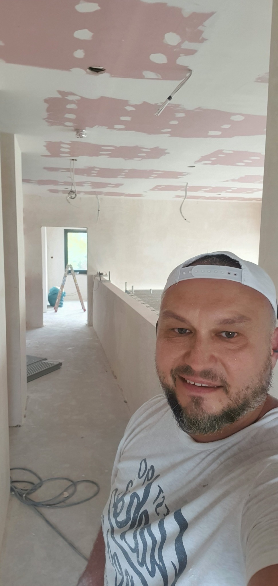 Mężczyzna w czapce robi selfie na tle wnętrza w trakcie szpachlowania sufitu, widoczne kable i drabina w oddali.