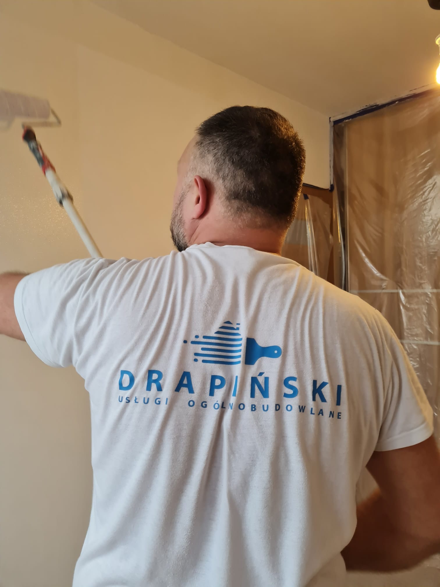 Mężczyzna maluje ścianę wałkiem, ubrany w białą koszulkę z logo firmy budowlanej DRAPIŃSKI USŁUGI OGÓLNOBUDOWLANE, zabezpieczone folią fragmenty pomieszczenia.