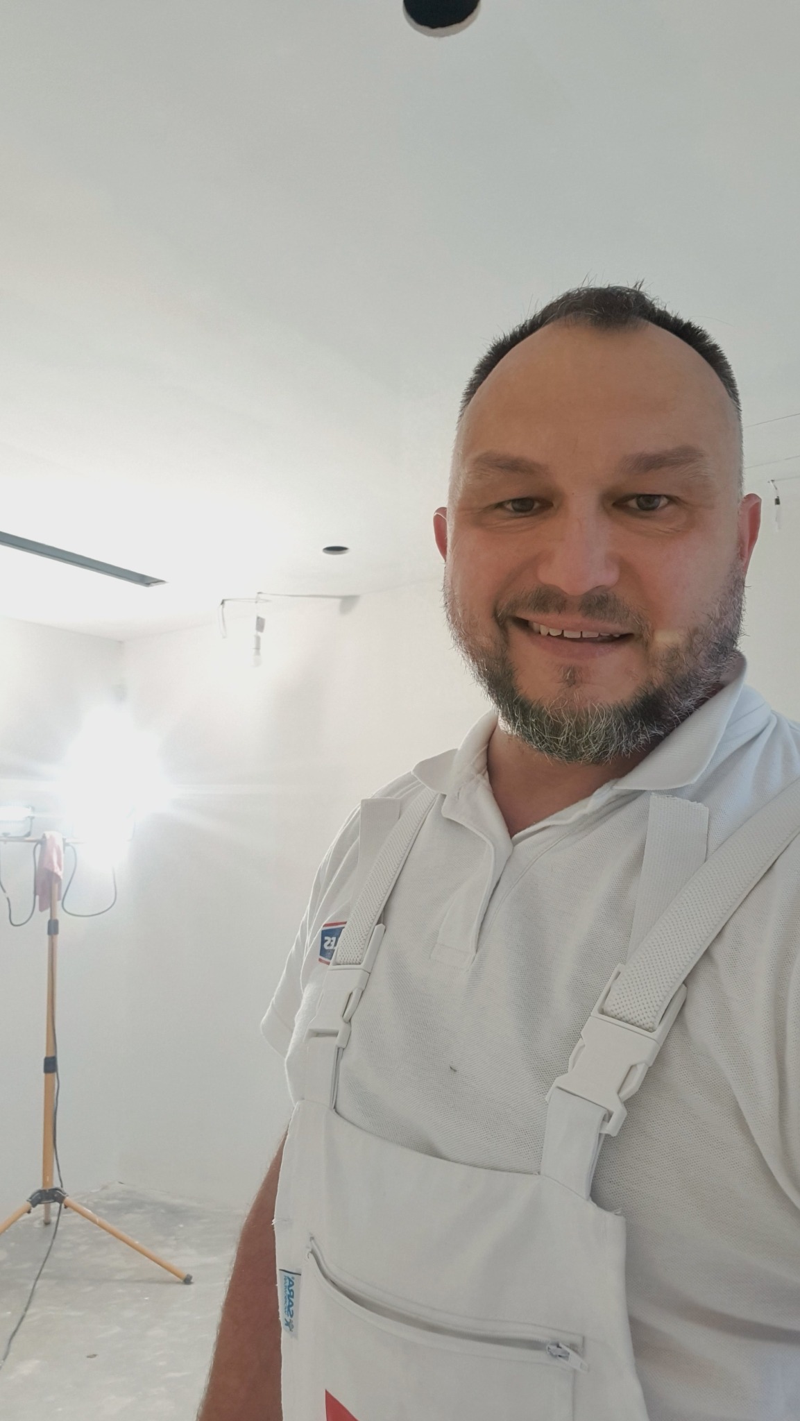 Uśmiechnięty malarz w białym kombinezonie na tle świeżo pomalowanego, jasnego pomieszczenia z widoczną lampą na statywie.