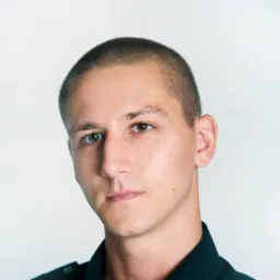 Portret młodego mężczyzny z krótkimi włosami, w ciemnej koszuli, na jasnym tle. Ujęcie z bliska, skupione na twarzy i wyrazie oczu.