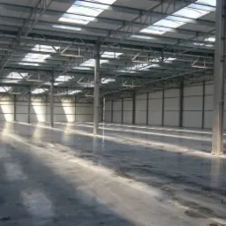 Wnętrze hali przemysłowej z betonową posadzką, widoczne metalowe słupy konstrukcyjne i świetliki w dachu, wpadające promienie słoneczne na posadzkę.