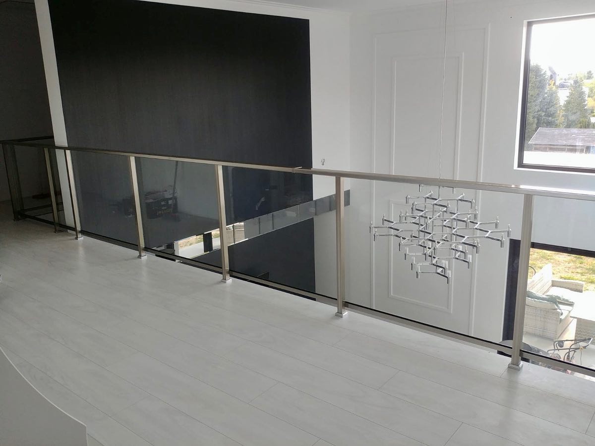 Wewnętrzna balustrada ze szkła dymionego i stali nierdzewnej na antresoli, widok z góry na białą podłogę, w tle czarna ściana i nowoczesny żyrandol.