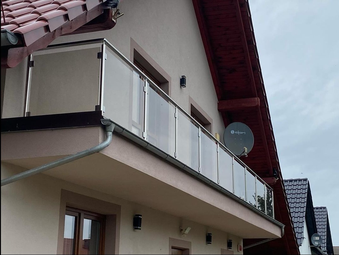 Balkon z przeszkloną balustradą ze stali nierdzewnej, widok z dołu, na tle domu z brązową dachówką i anteną satelitarną.