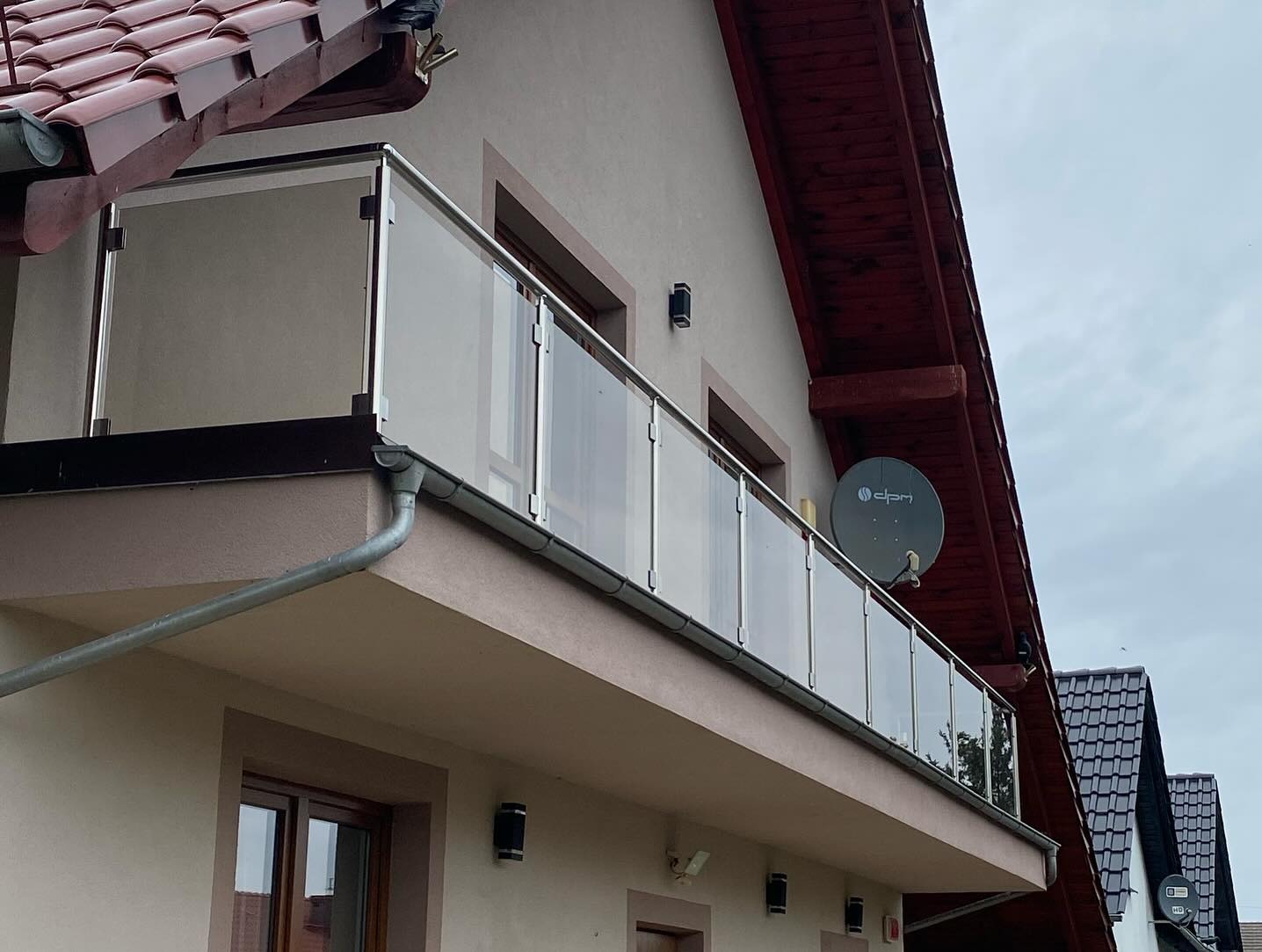 Balkon z nowoczesną, szklaną balustradą z metalowymi słupkami i poręczą, zamontowany w budynku z beżową elewacją i brązowym dachem.