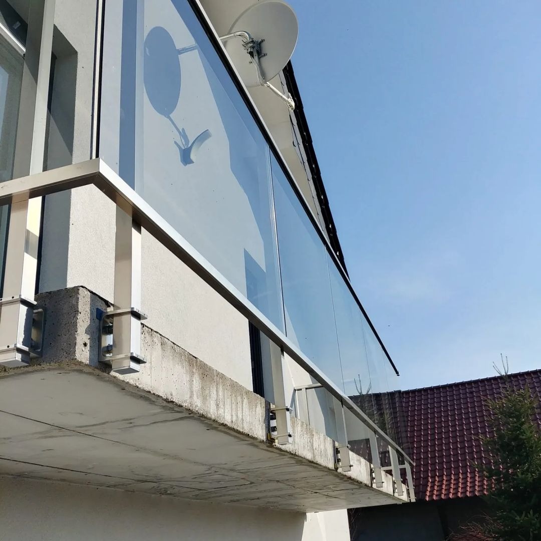 Szklana balustrada balkonowa z metalowymi słupkami, mocowana do betonowej płyty balkonu, widok z dołu na tle błękitnego nieba i fragmentu dachu sąsiedniego budynku.