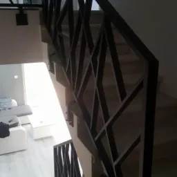 Nowoczesna, czarna balustrada schodowa o geometrycznym wzorze, widok z góry na jasne schody i salon z białymi meblami.