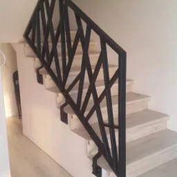 Nowoczesna, czarna balustrada schodowa z geometrycznym wzorem na jasnych, drewnopodobnych schodach i podłodze, widok z dołu.