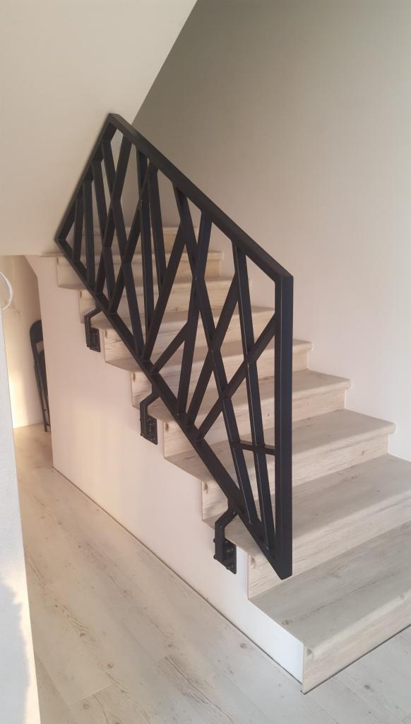Nowoczesna, czarna balustrada schodowa z geometrycznym wzorem na jasnych, drewnopodobnych schodach i podłodze, widok z dołu.