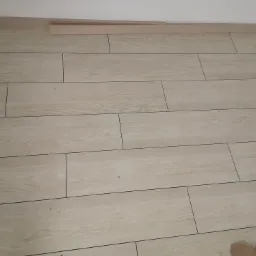 Ułożona podłoga z płytek imitujących drewno w odcieniach beżu, z widoczną listwą przypodłogową i fragmentami kartonu w rogu.