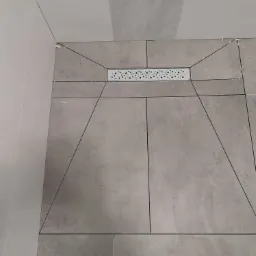 Ułożone szare płytki podłogowe w kabinie prysznicowej z odpływem liniowym, widoczne krzyżyki dystansowe.