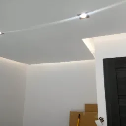 Nowoczesny sufit podwieszany z oświetleniem LED, wpuszczone lampy punktowe, świeżo pomalowane ściany, widoczny fragment ciemnych drzwi i kartony na podłodze.