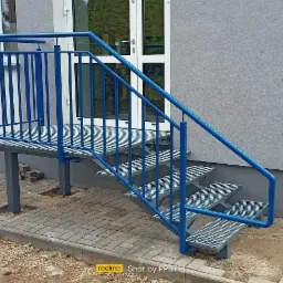 Zewnętrzne metalowe schody z niebieską balustradą prowadzące do budynku, kostka brukowa w trakcie układania.