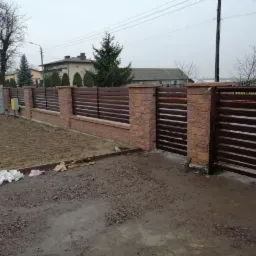 Nowoczesne ogrodzenie z brązowymi, poziomymi przęsłami i słupkami z beżowej cegły klinkierowej, zakończone bramą wjazdową. Widok z zewnątrz posesji.