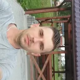 Mężczyzna na tle drewnianej altany z szarą dachówką. Widoczne elementy konstrukcyjne i fragment posesji. Ujęcie z bliska, lekko z dołu. T-shirt z napisem.