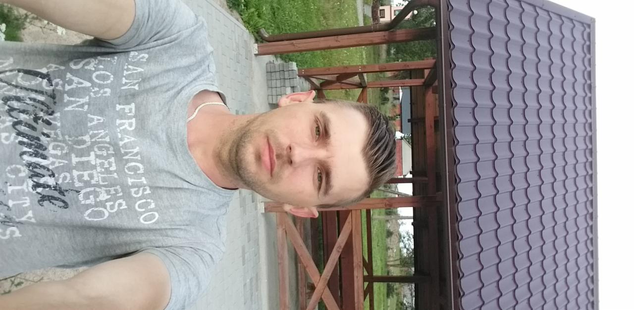 Mężczyzna na tle drewnianej altany z szarą dachówką. Widoczne elementy konstrukcyjne i fragment posesji. Ujęcie z bliska, lekko z dołu. T-shirt z napisem.