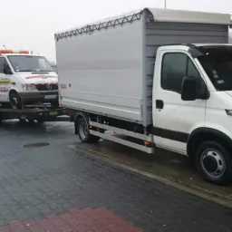 Biała laweta z pomarańczowym światłem ostrzegawczym przewożona na platformie przez inny biały samochód dostawczy z plandeką, stojący na stacji benzynowej.