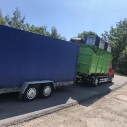 Ciężarówka z zielonym kontenerem wypełnionym odpadami, ciągnąca niebieską przyczepę z plandeką, na tle drzew i błękitnego nieba.