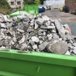 Zielony kontener wypełniony gruzem betonowym i ceglanym, widoczny na tle budynków i innych kontenerów.