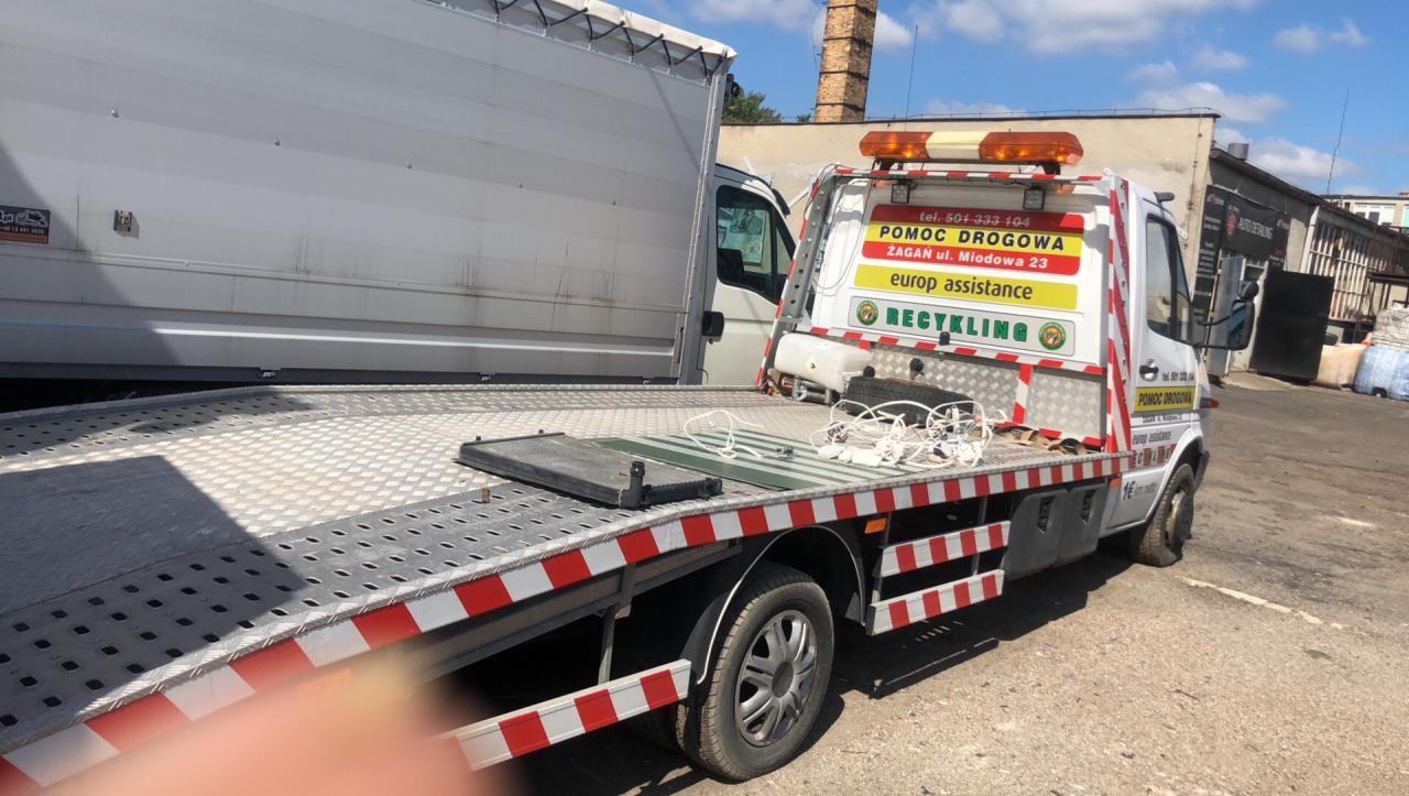 Laweta z platformą załadowczą, częściowo załadowana białym busem, z widocznym napisem 'Pomoc Drogowa Żagań' i numerem telefonu, zaparkowana przed budynkiem z ceglanym kominem.