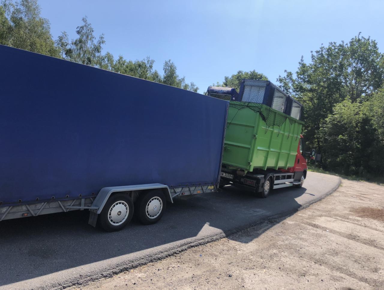 Ciężarówka z zielonym kontenerem wypełnionym odpadami, ciągnąca niebieską przyczepę z plandeką, na tle drzew i błękitnego nieba.