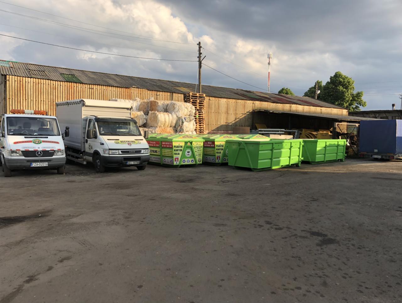 Dwa busy transportowe i kilka zielonych kontenerów na odpady ustawione na placu przed magazynem z belami makulatury w tle.
