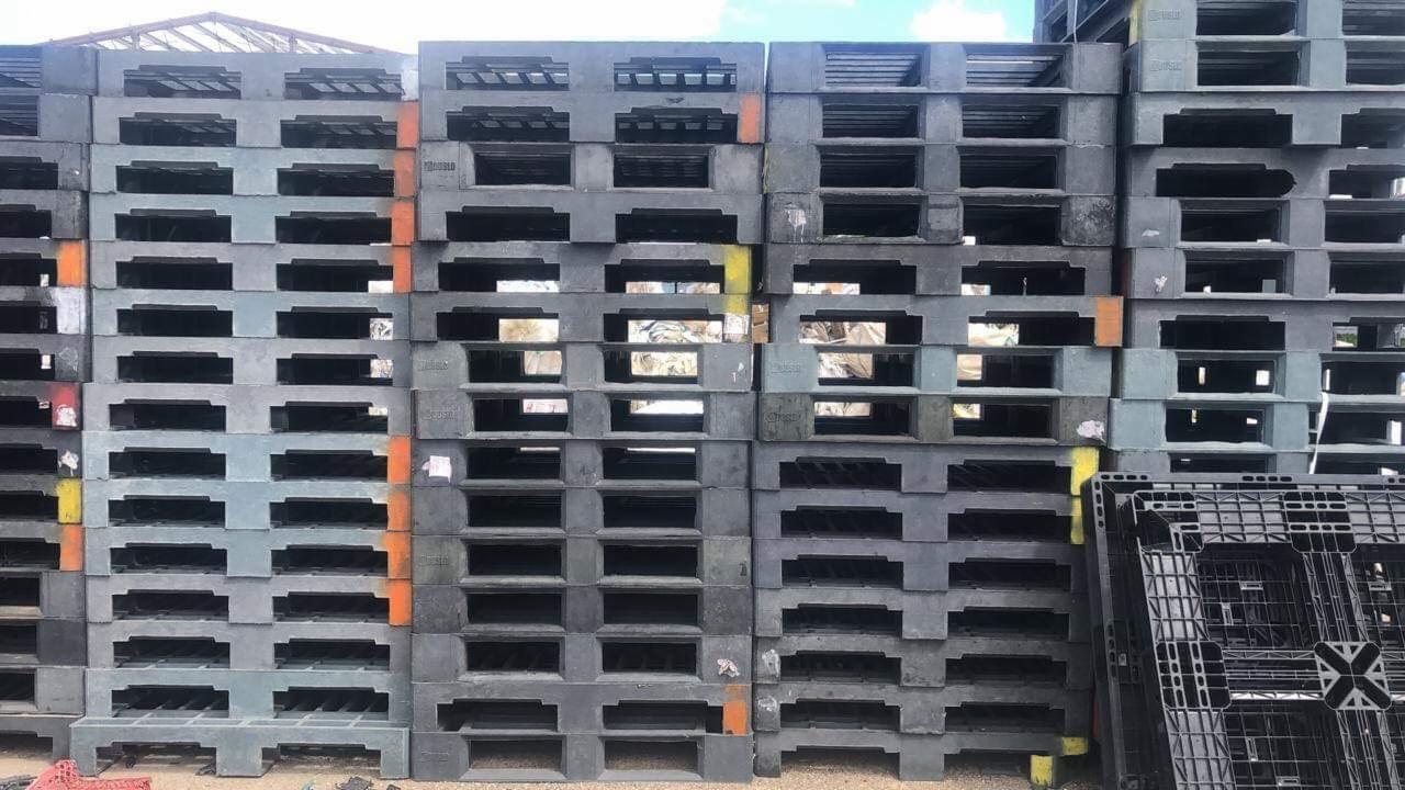 Stos szarych plastikowych palet transportowych z pomarańczowymi i żółtymi oznaczeniami, ustawionych na zewnątrz pod zadaszeniem.