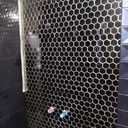 Ściana w łazience wyłożona czarnymi heksagonalnymi płytkami z białymi fugami, widoczne wystające rurki instalacji wodnej i kable elektryczne przed ostatecznym wykończeniem.