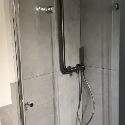 Nowoczesna kabina prysznicowa z czarną armaturą i szklanymi drzwiami, wykończona szarą płytką imitującą beton. Minimalistyczny design z odpływem liniowym.