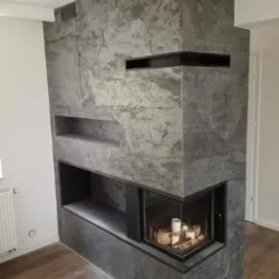 Nowoczesny kominek obłożony szarym kamieniem z czarnymi wnękami, widoczne drewno i świece w palenisku, podłoga drewniana.