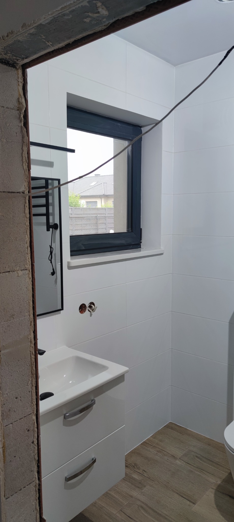 Nowoczesna, jasna łazienka w trakcie wykańczania: białe płytki na ścianach, drewnopodobne panele na podłodze, biała szafka pod umywalką i okno z widokiem na dom.