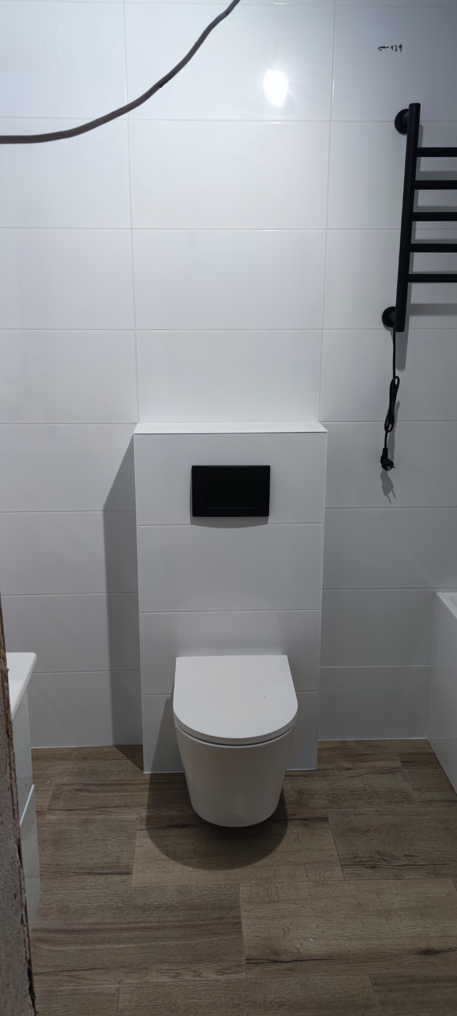 Nowoczesna toaleta podwieszana z czarnym przyciskiem spłukującym na tle białych płytek i drewnopodobnej podłogi, uzupełnona czarnym grzejnikiem.