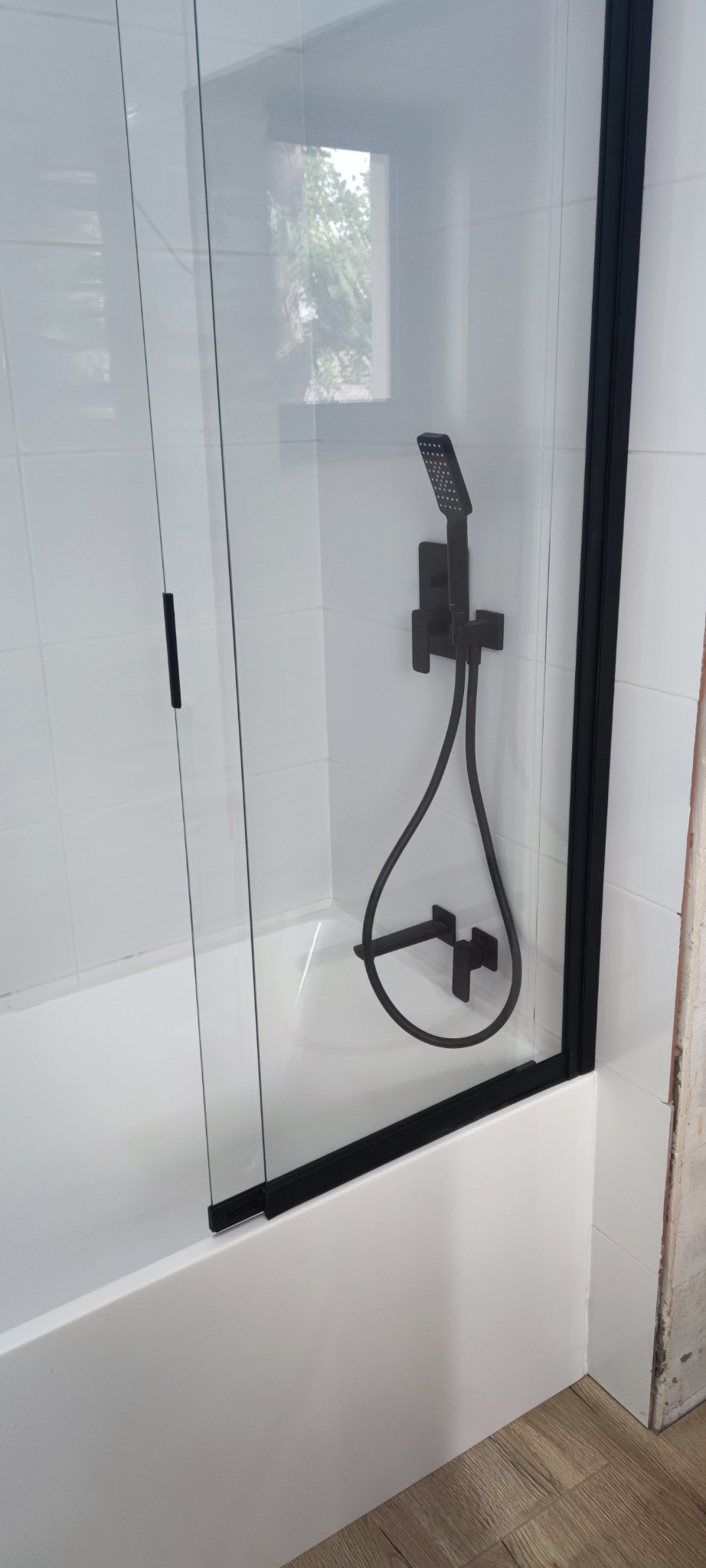 Nowoczesna kabina prysznicowa z czarnymi elementami, białe płytki, widok na okno z zielenią. Minimalistyczny design, świeżo po remoncie.