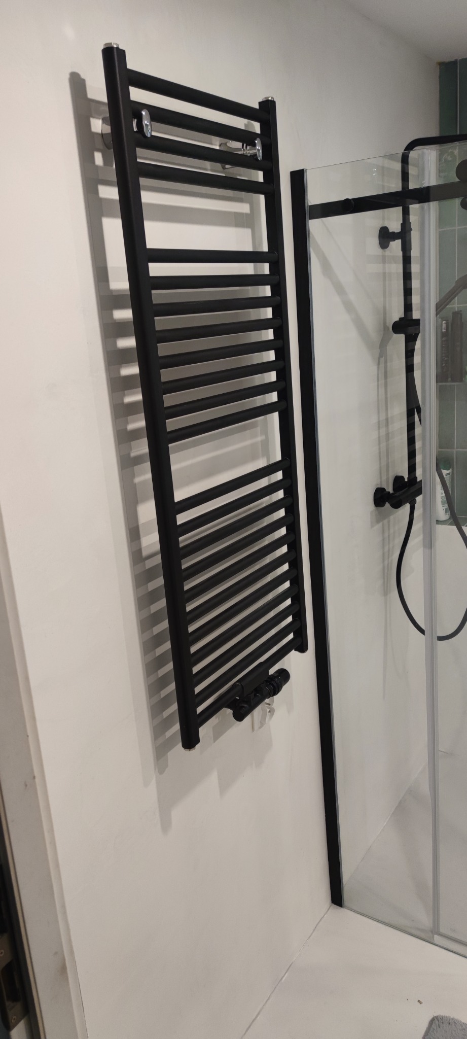 Czarny, drabinkowy grzejnik łazienkowy na białej ścianie, obok kabiny prysznicowej z czarnymi elementami. Widoczne chromowane wieszaki na grzejniku.