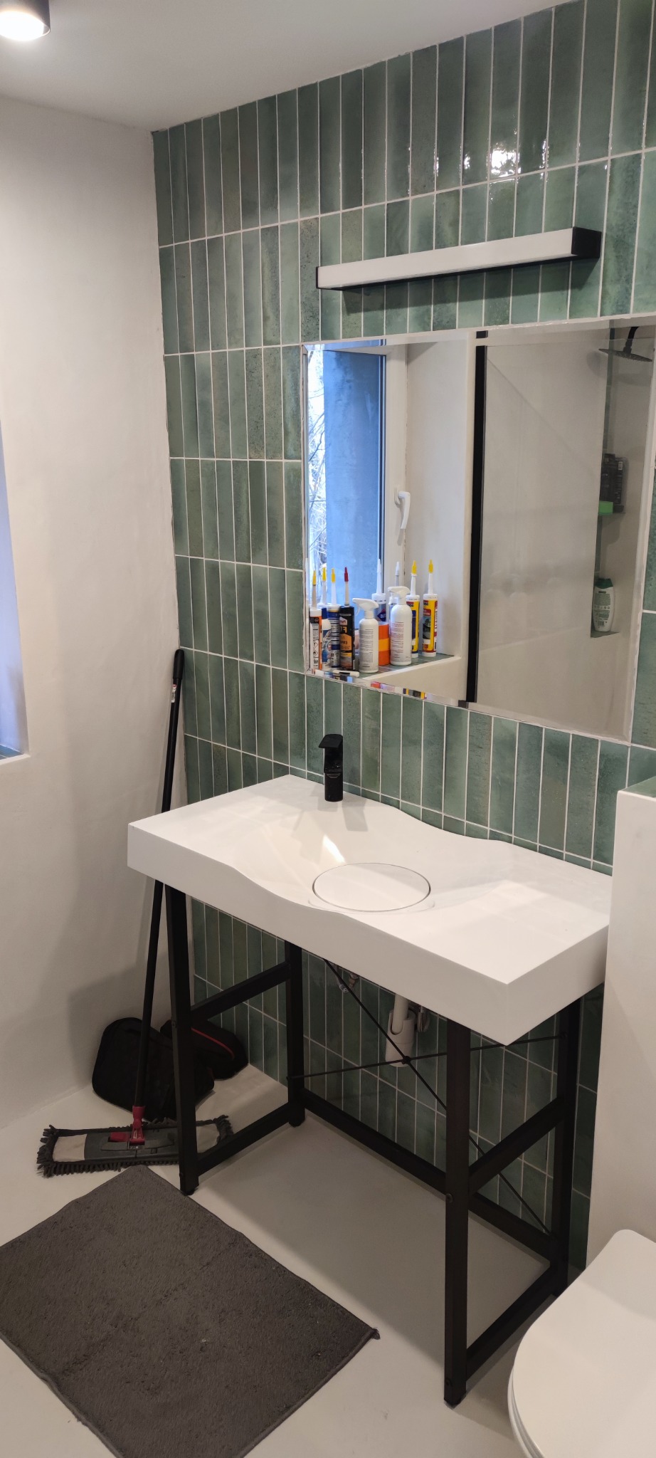 Wykończona łazienka z umywalką na czarnym stelażu, zielone płytki na ścianie i czarna bateria. W lustrze odbijają się tuby z silikonem i inne akcesoria.