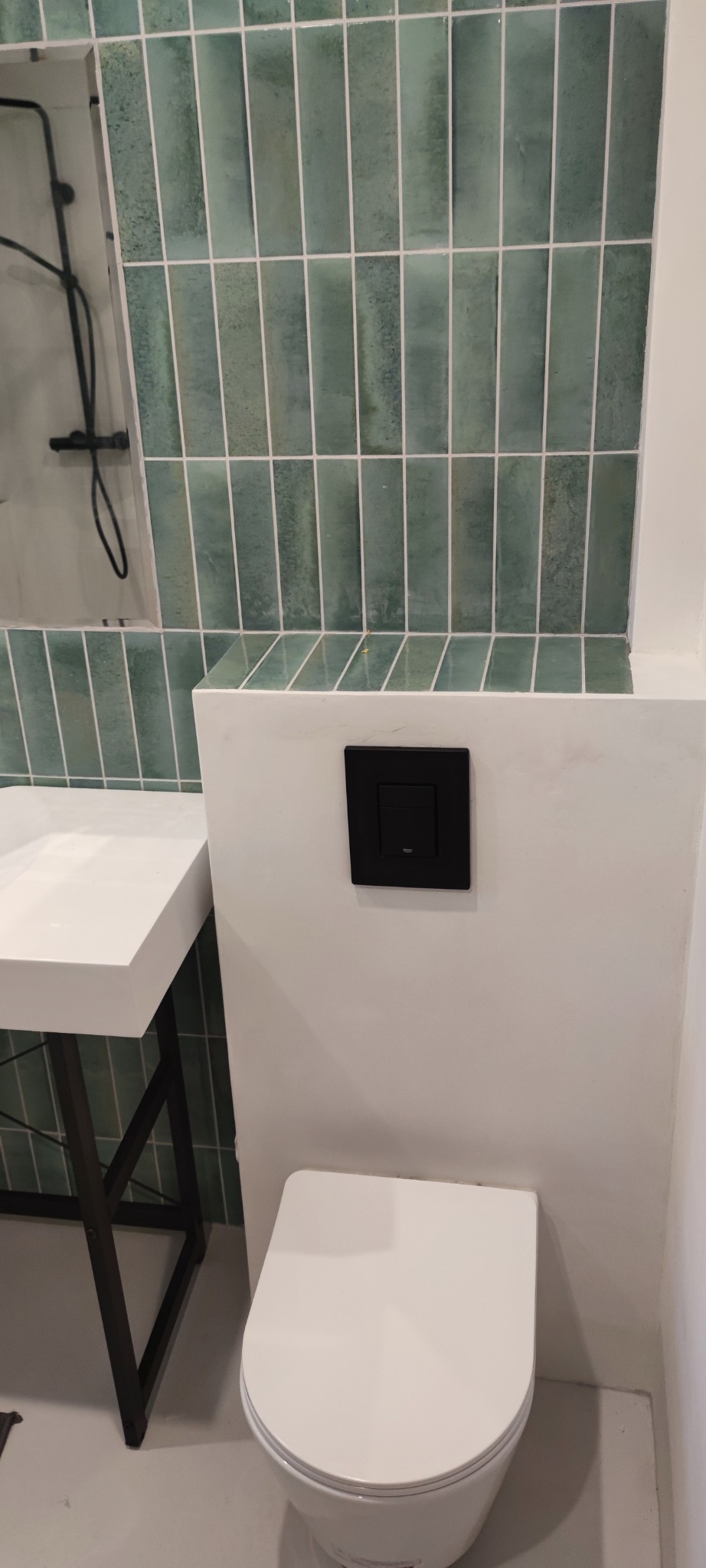 Nowoczesna łazienka: biała toaleta podwieszana, umywalka na metalowym stelażu i ściana z zielonej, wąskiej glazury. Czarny przycisk spłuczki.