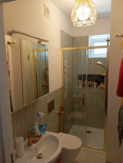 Wykończona łazienka z kabiną prysznicową w złotej ramie, umywalką i toaletą. Ściany wyłożone płytkami w odcieniach zieleni. W tle widoczne okno.