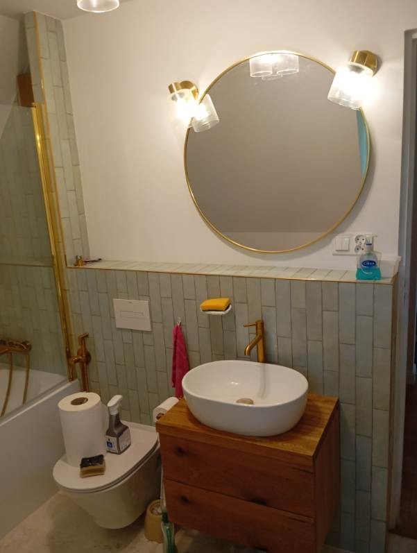 Wykończona łazienka z okrągłym lustrem w złotej ramie, drewnianą szafką pod umywalką i płytkami w odcieniach szarości. Uwagę przykuwają złote akcenty.
