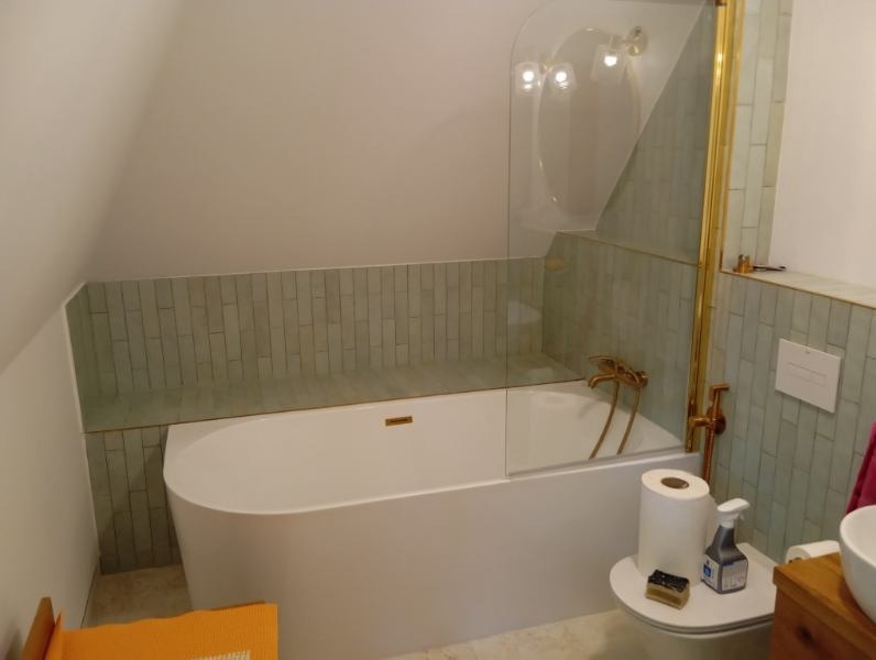 Nowoczesna łazienka na poddaszu z białą wanną, złotą armaturą i zieloną glazurą ułożoną pionowo. Kabina prysznicowa ze szkła.