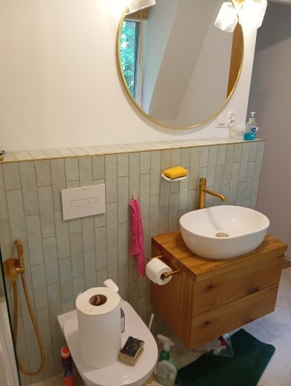 Jasna łazienka z okrągłym lustrem w złotej ramie, drewnianą szafką pod umywalką i toaletą z zapasem papieru. Minimalistyczny design, złote akcenty.