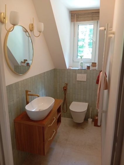 Nowoczesna łazienka na poddaszu z umywalką nablatową, złotą armaturą i jasnymi płytkami, doświetlona oknem z żaluzjami. Minimalistyczny design.