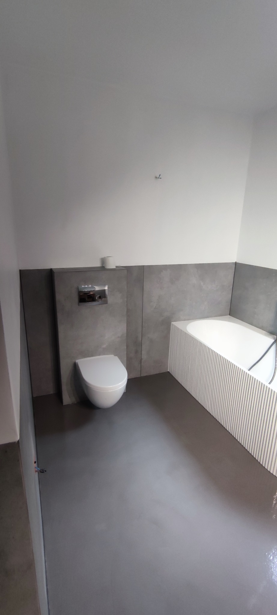 Nowoczesna łazienka z szarą podłogą i panelami, białą wiszącą toaletą i wanną z pionowymi żłobieniami. Minimalistyczny design, widok na fragment wnętrza.