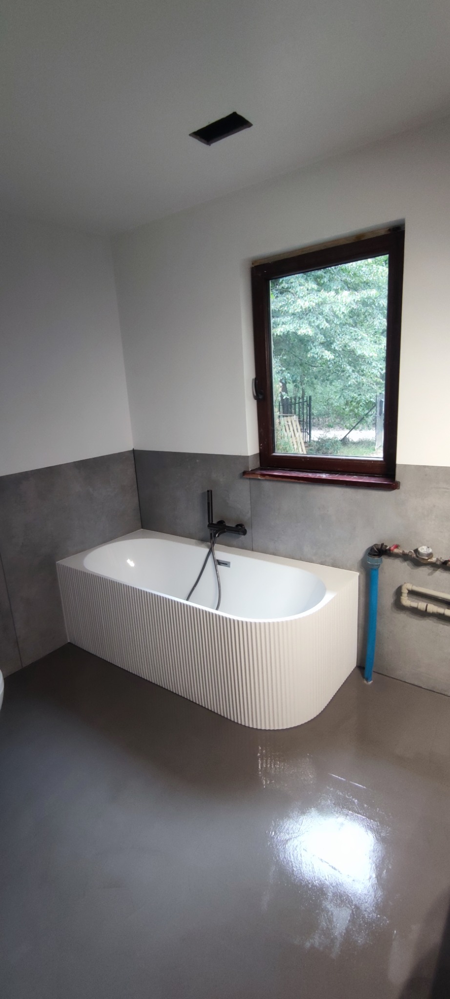 Nowoczesna łazienka z białą wanną o żłobkowanej obudowie, szarą posadzką żywiczną i oknem z widokiem na zieleń. Minimalistyczny design, eleganckie wykończenie.