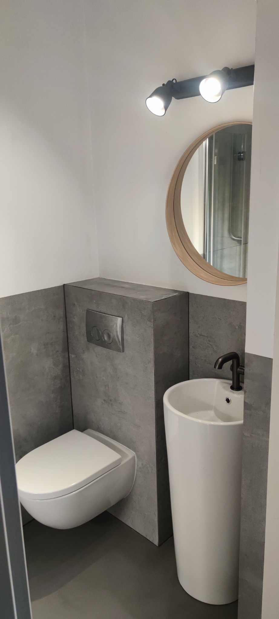 Nowoczesna łazienka z podwieszaną toaletą i umywalką wolnostojącą. Szare płytki imitujące beton na ścianach, okrągłe lustro z drewnianą ramą i czarna bateria umywalkowa.