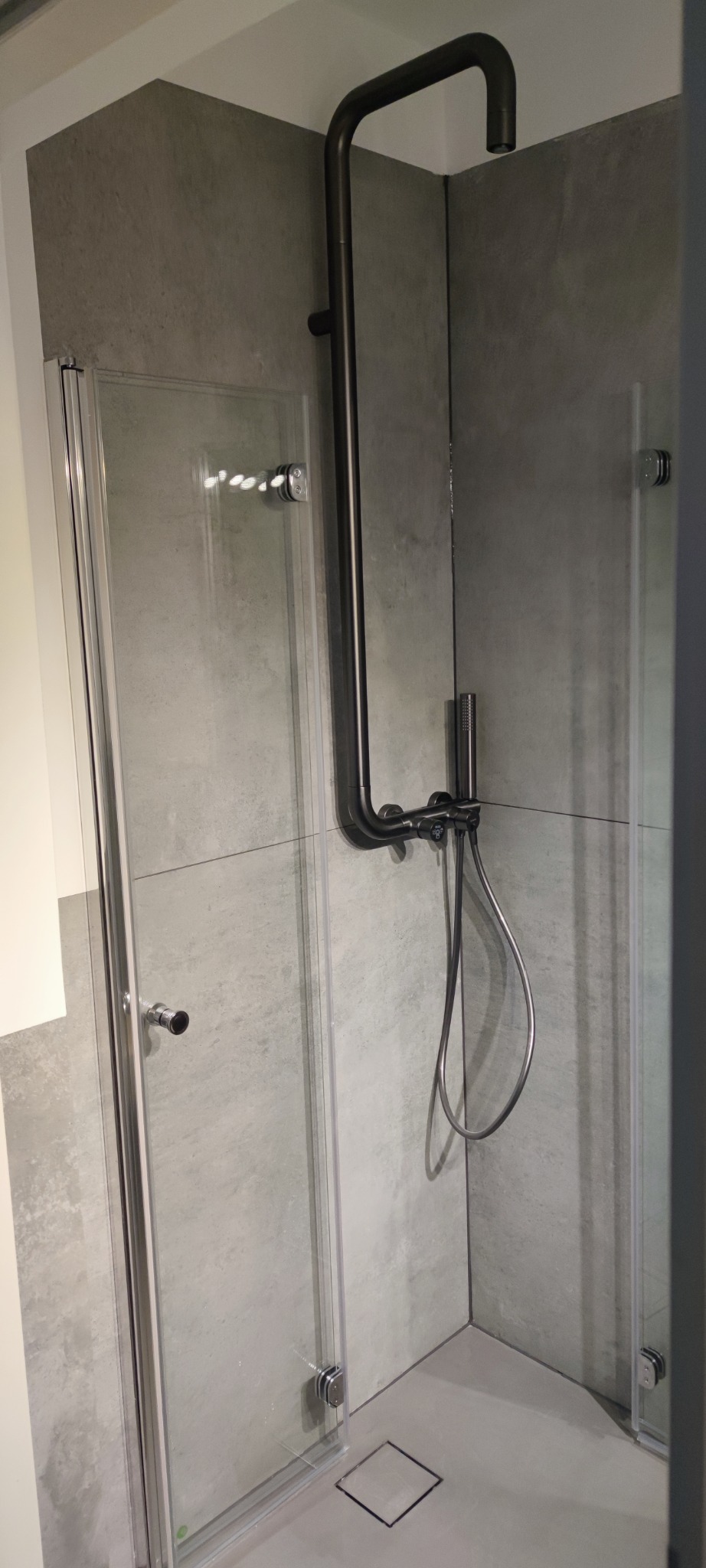 Nowoczesna kabina prysznicowa z czarną armaturą i szklanymi drzwiami, wykończona szarą płytką imitującą beton. Minimalistyczny design z odpływem liniowym.